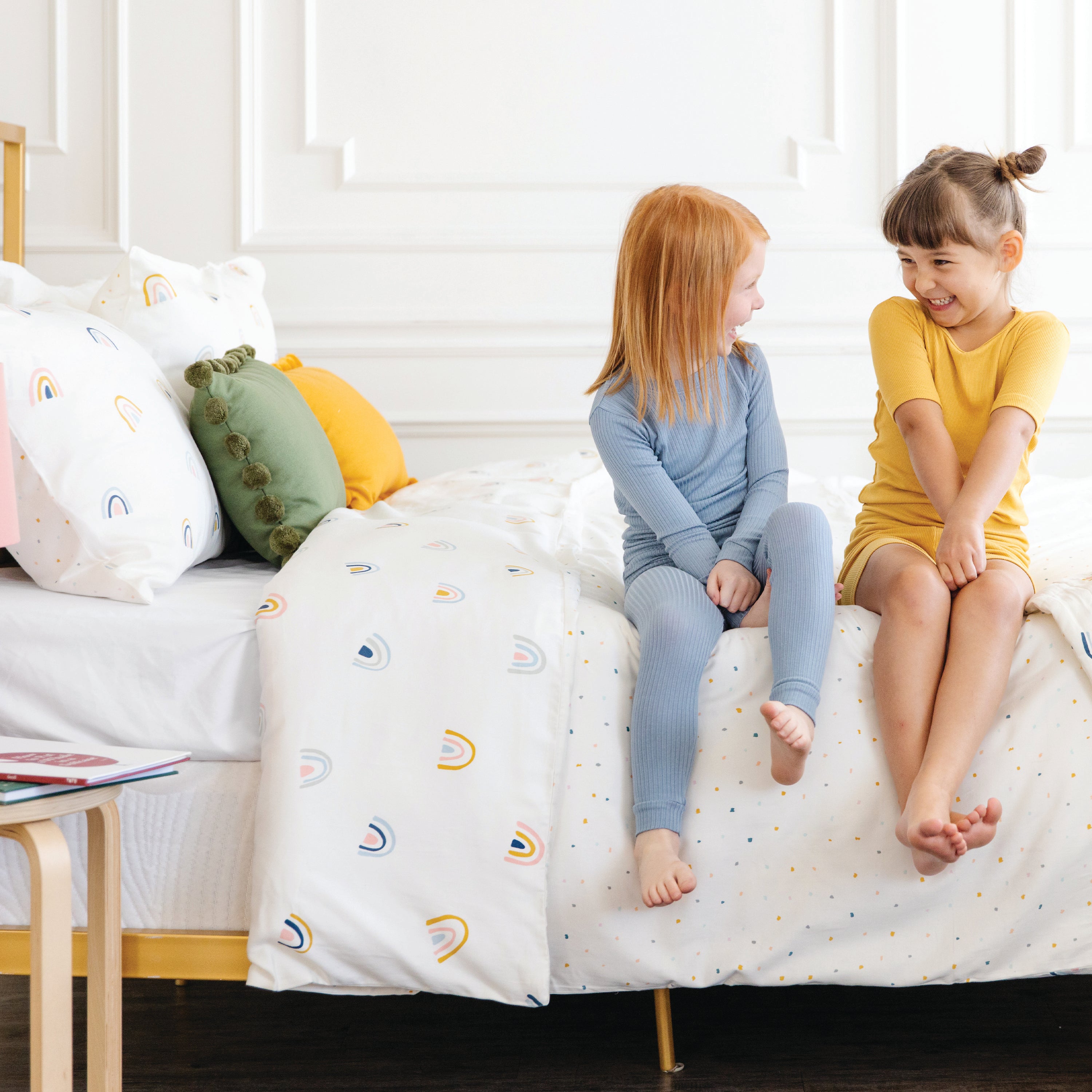 Two young girls laughing and chatting while sitting on a white bed with the Makemake Organics Organic Duvet Cover - Dotty & Rainbow bedding in a bright, cozy room.