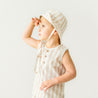 A young toddler wearing a Makemake Organics Organic Linen Bucket Sun Hat - Beige Stripes and matching tank top looks into the distance with a focused expression, standing against a plain, light-colored background.