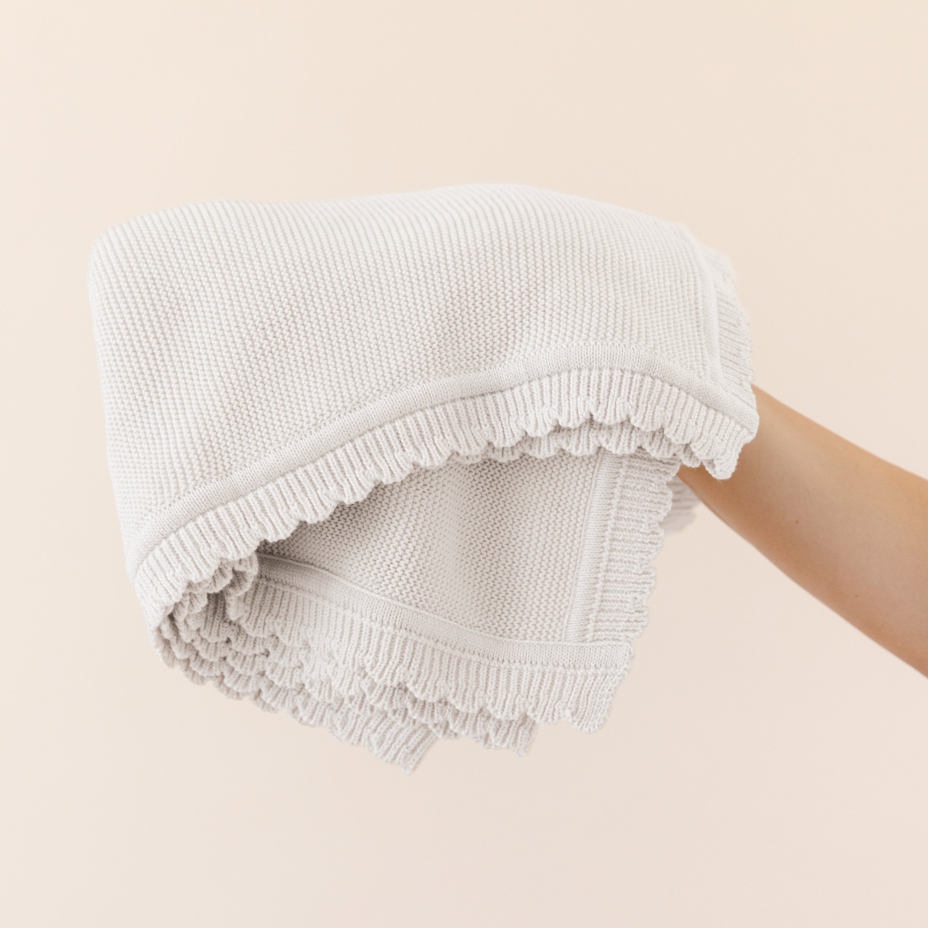 A person holding a folded Organic Cotton Scalloped Baby Blanket - Ella Ivory by Makemake Organics with a textured pattern and fringed edges against a soft beige background.