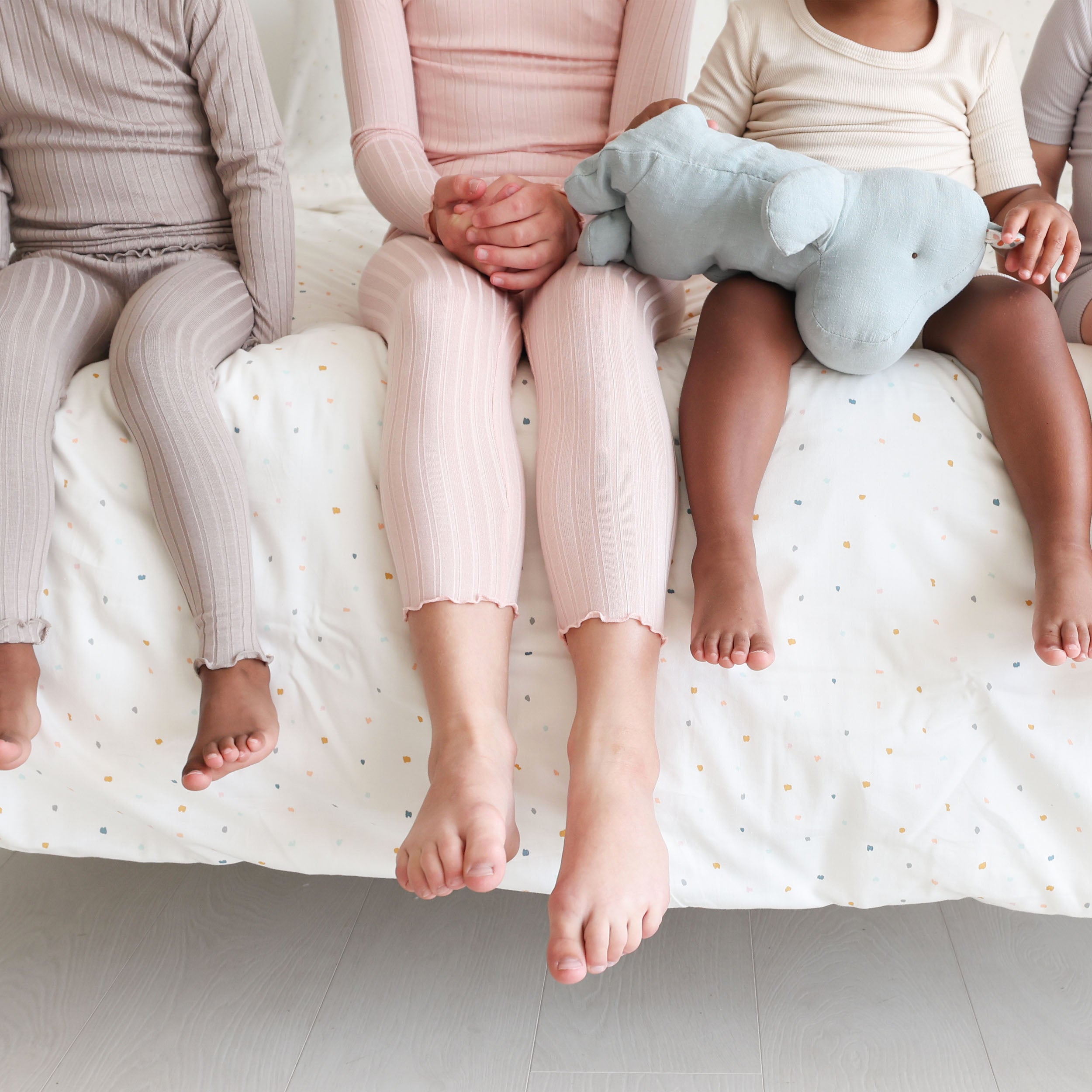 Three children sit side by side on a bed, showing only their lower bodies. they wear pajamas; the middle child holds a blue elephant plush toy. the bedspread is white with colorful dots using the Makemake Organics Organic Cotton Sheet Set - Dotty.