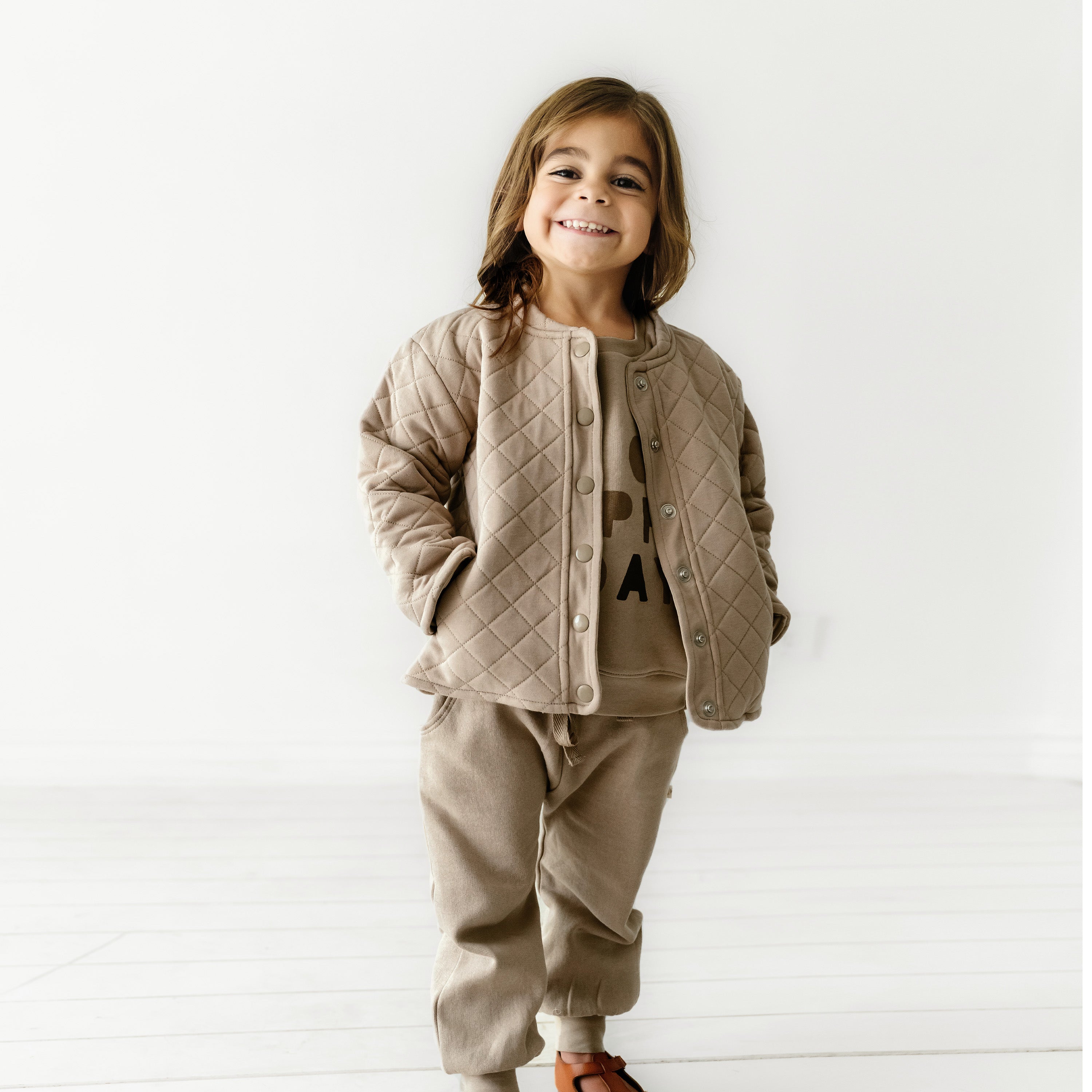A joyful young girl stands in a well-lit room, wearing an Organic Kids organic merino wool buttoned jacket in mocha and matching pants, with a playful smile on her face.