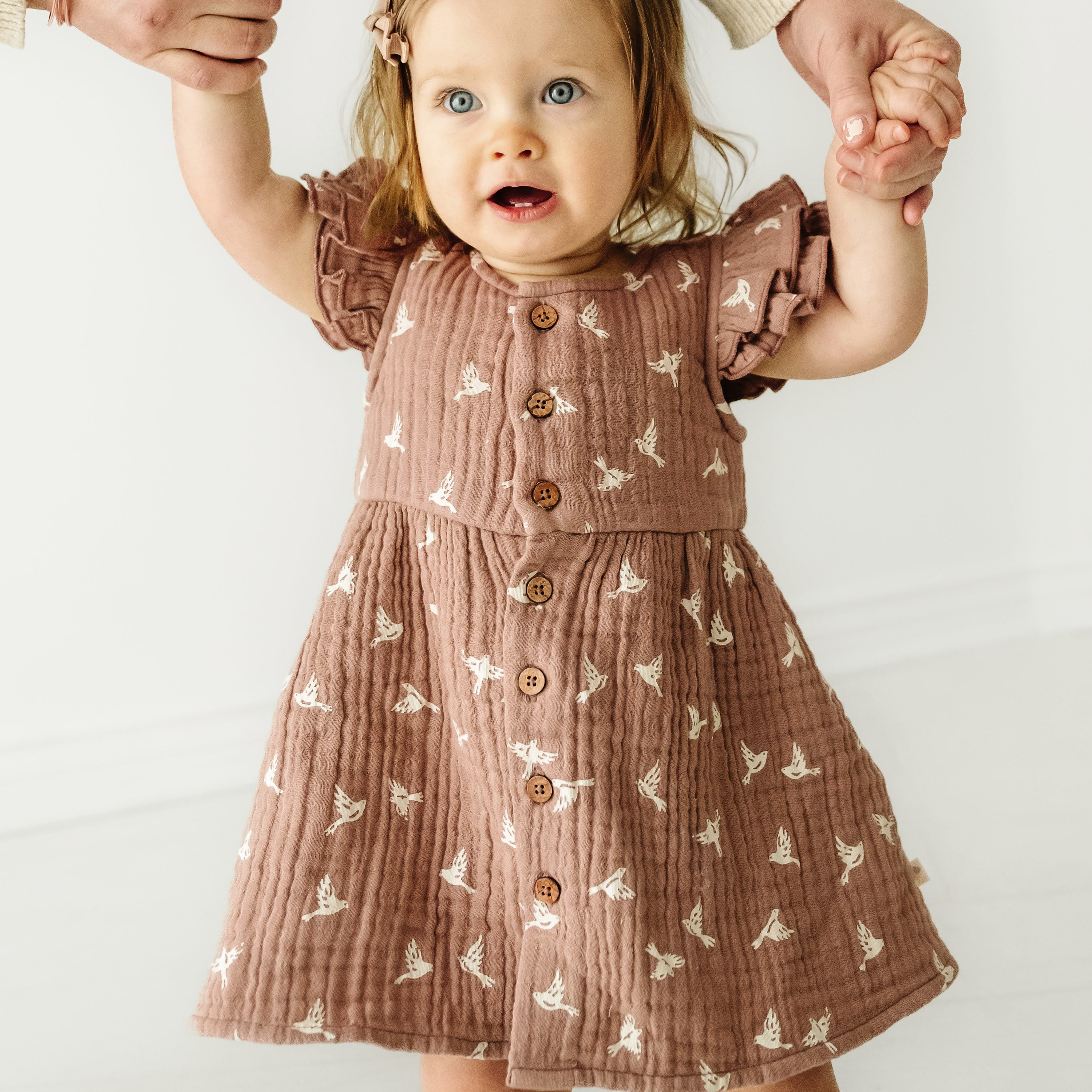 A toddler girl in a Organic Muslin Button Flutter Dress - Flock by Makemake Organics looks up with wide eyes and excitement, holding an adult’s hands. She stands against a clean, white background.