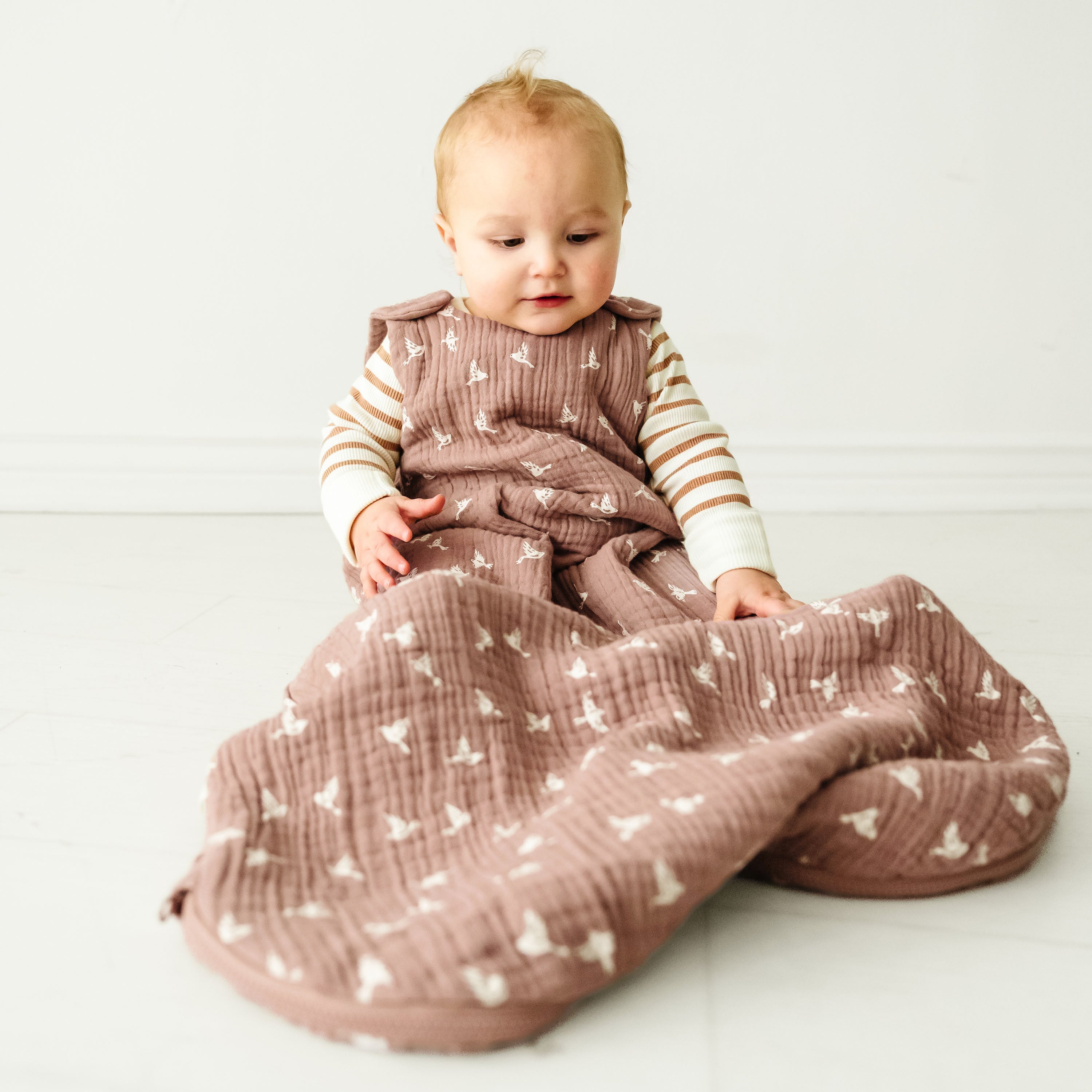 A toddler with light hair sits on a white floor, wearing a striped long-sleeve shirt and a puffy sleeveless jumper, holding a Makemake Organics Muslin Wearable Blanket - Flock with a geometric pattern.