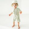 A toddler girl in a green and white checkered romper and Makemake Organics Organic Muslin Bucket Sun Hat - Gingham stands barefoot on a white floor, looking directly at the camera with a curious expression.