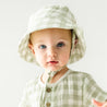 A baby with blue eyes wearing a light green checkered outfit and an Organic Muslin Bucket Sun Hat in Gingham by Makemake Organics, looking directly at the camera with a subtle expression.