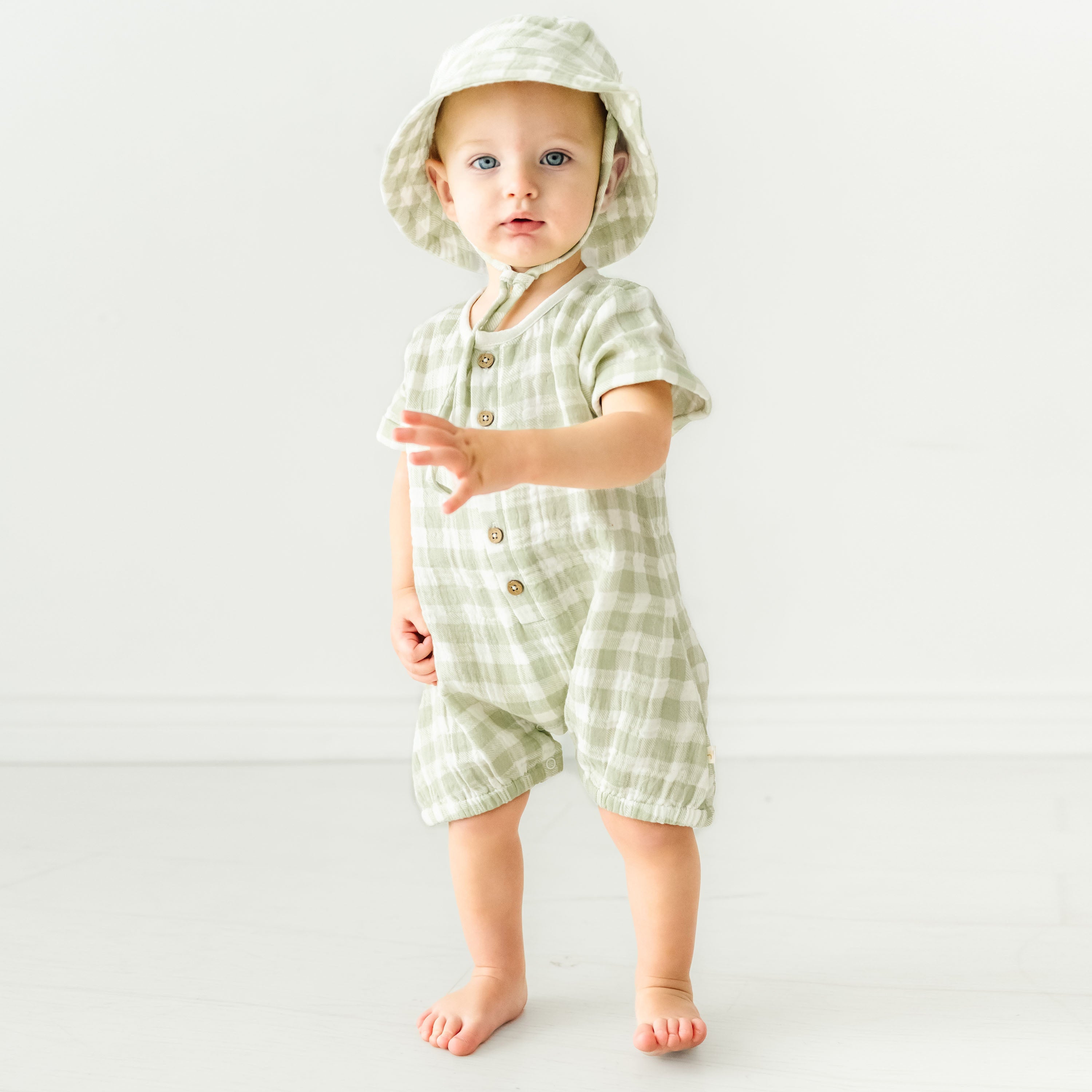 A toddler girl wearing a Makemake Organics Organic Muslin Short Bubble Romper - Gingham and matching sun hat stands on a white background, looking curiously at the camera.