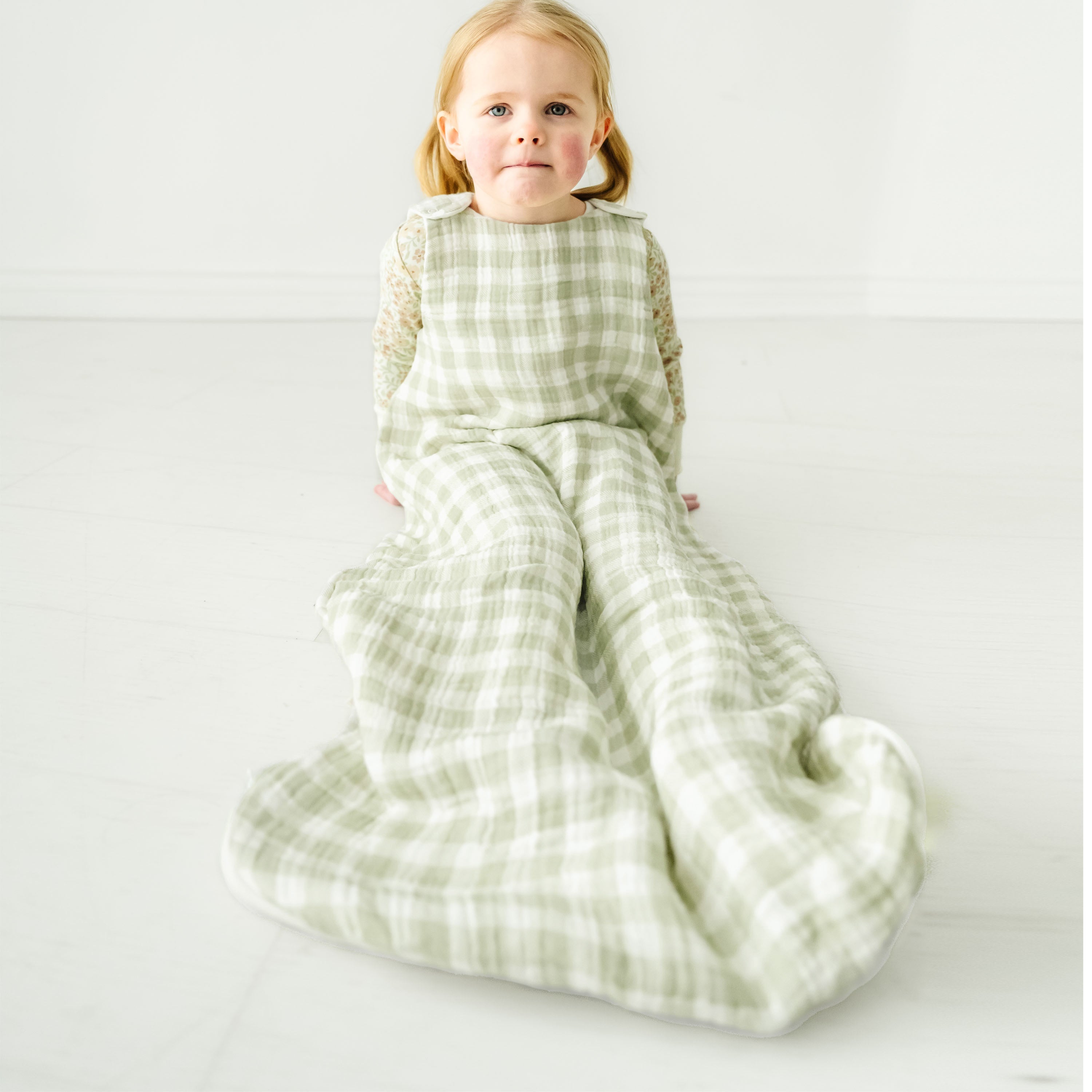 A young toddler with blonde hair sits on the floor wearing a Makemake Organics Muslin Wearable Blanket - Gingham. The child's expression is calm and gentle, against a soft white background.