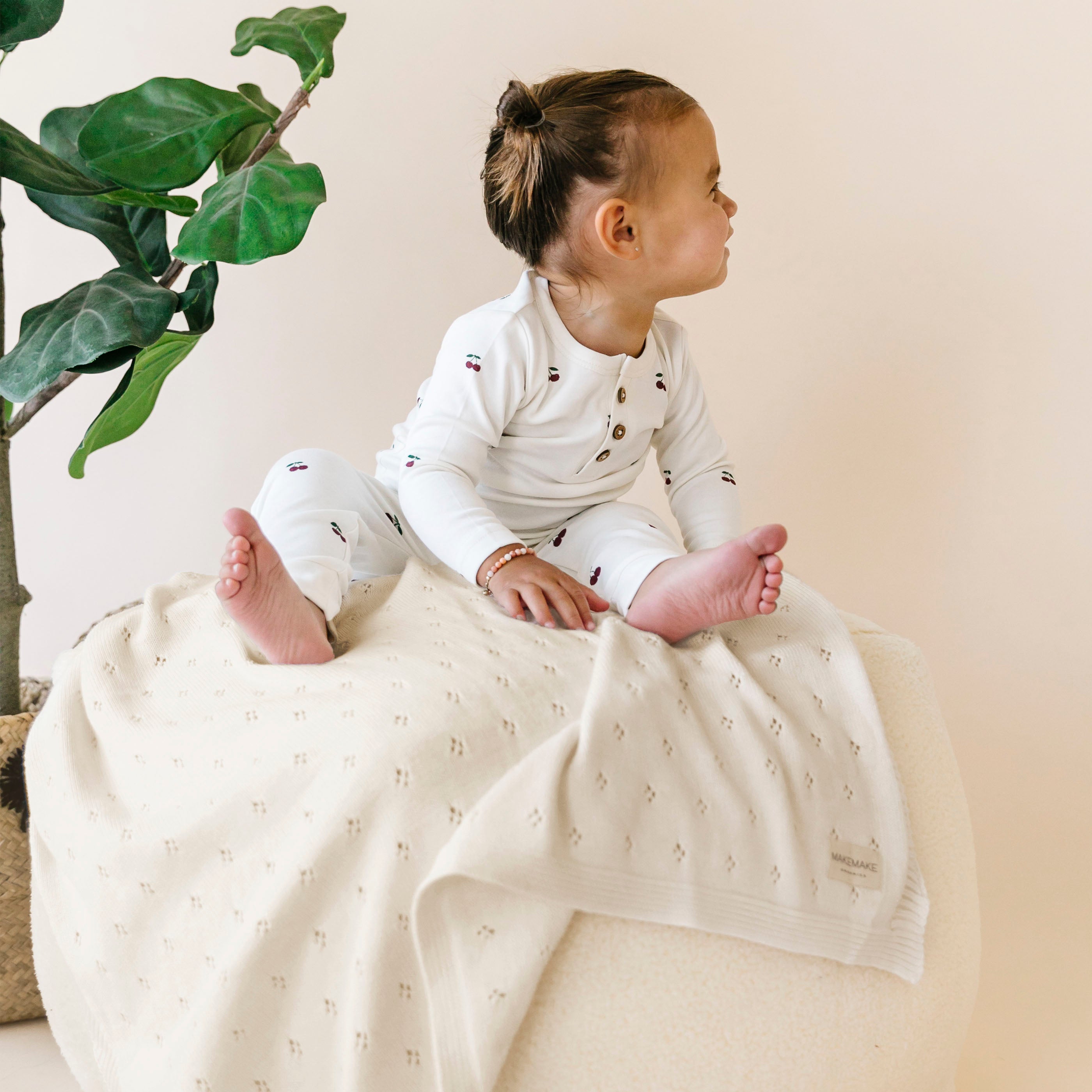 Toddler dressed in a white patterned onesie sitting on a Makemake Organics Organic Cotton Pointelle Baby Blanket - Vanilla Natural over a pouf, with a green plant in the background. the child has their hair tied up in a bun.