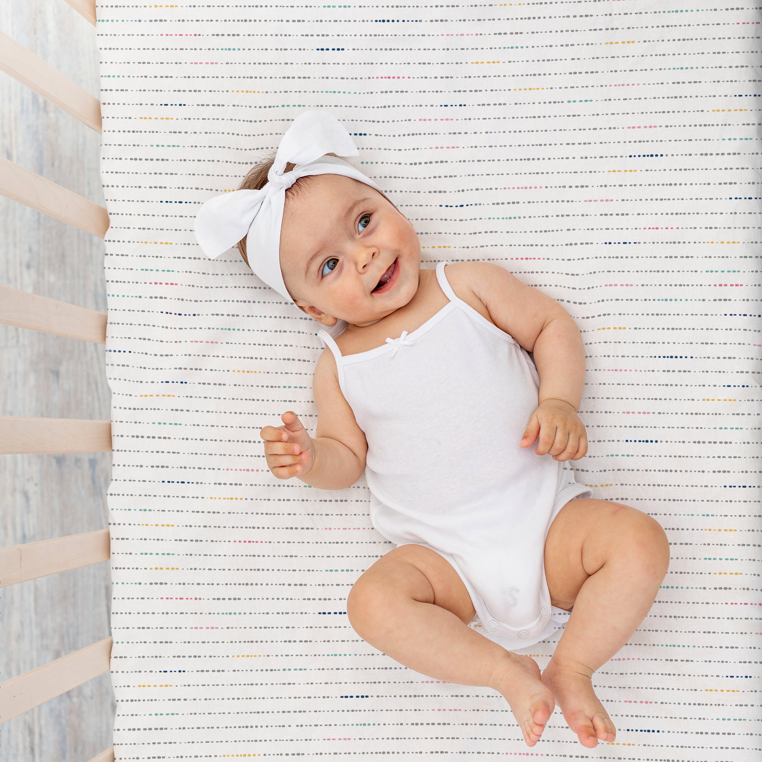 A happy baby wearing a white onesie and a matching bow headband, lying on a striped gray and white mat, smiling and looking upward on the Makemake Organics Mini Crib Fitted Sheet in Pebble Pop.