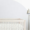 A clean, minimalist baby room with a white Celestial crib adorned with a patterned Makemake Organics bedding set. A floor lamp with a metallic finish stands beside it, all against a light gray wall with a subtle round paint detail.