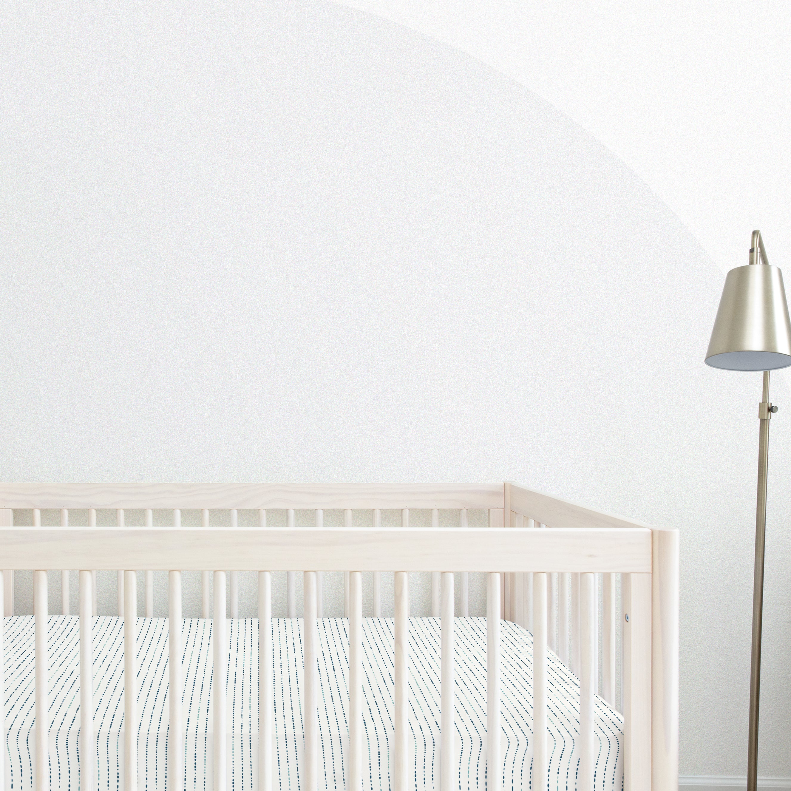 A minimalist nursery with a white Makemake Organics crib fitted sheet with pillowcase against a white wall with a large, partial grey circle painted on it. a metallic floor lamp stands to the right.