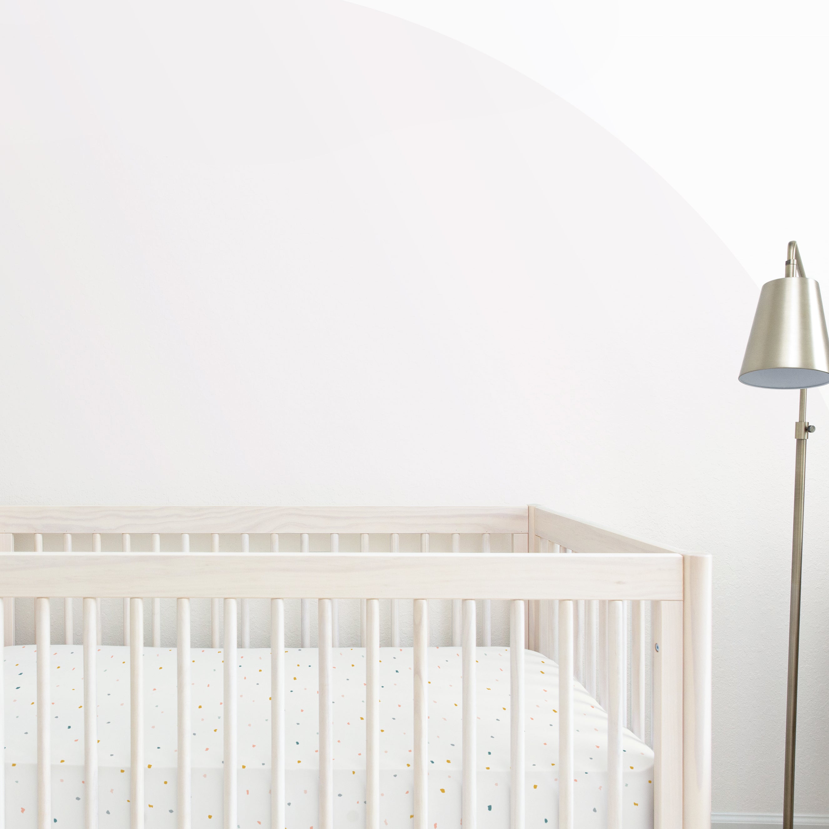 A minimalist nursery room with a white crib featuring the Makemake Organics Crib Fitted Sheet with Pillowcase - Dotty bedding and a standing lamp with a white shade. the walls are soft white with a subtle, large pastel arch design.