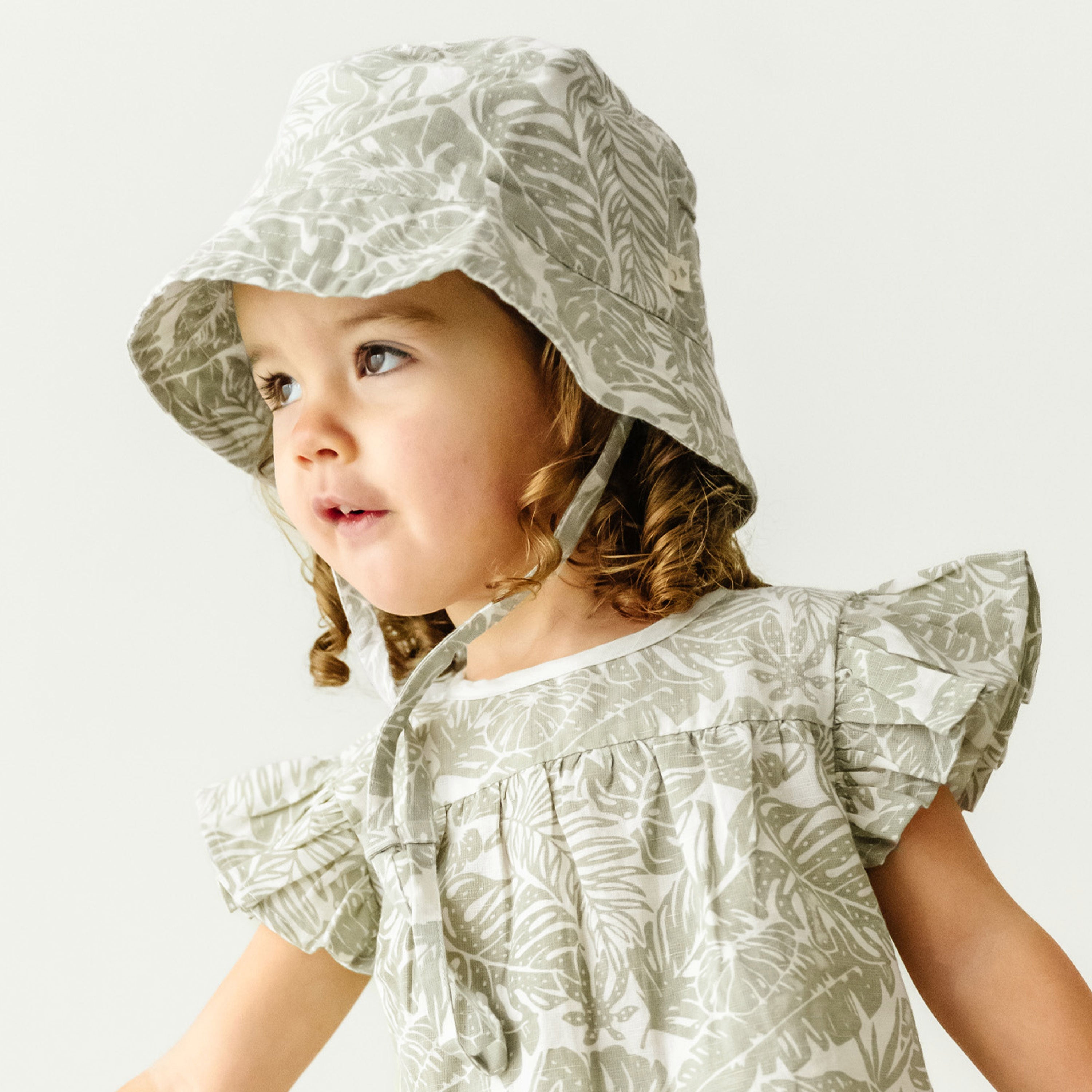 A toddler with braided hair, wearing a patterned dress and the Makemake Organics Organic Linen Bucket Sun Hat - Palms, looks to the side with a curious expression on a plain light background.
