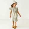 Sentence with replaced product:
"A baby wearing a patterned outfit with ruffled sleeves and a Makemake Organics Organic Linen Bucket Sun Hat - Palms, paired with tan shoes, standing in a white studio.