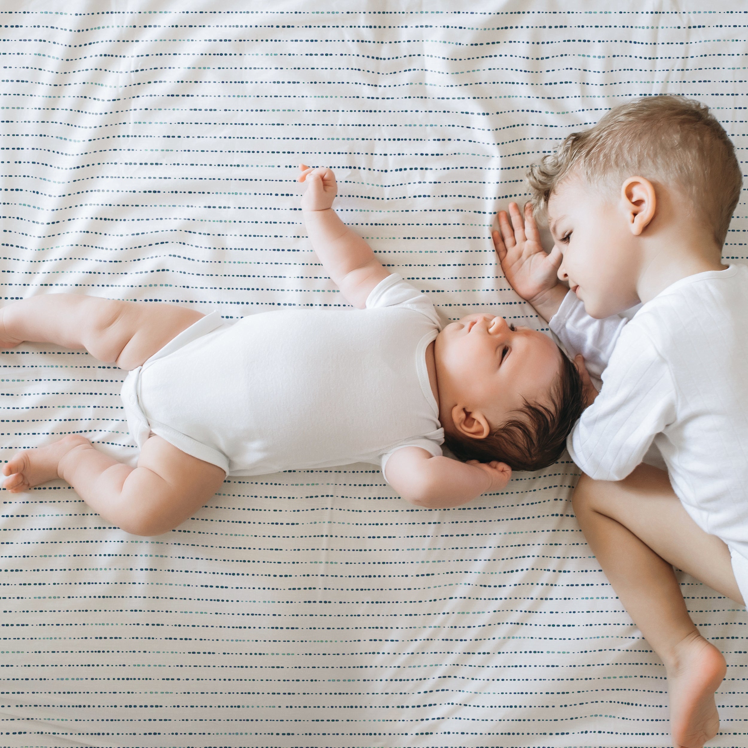 Two young children lying on a Makemake Organics Organic Cotton Sheet Set - Pebble. the older child, wearing a white shirt, gently touches the younger child's head, who is in a white onesie, stretching an arm out.