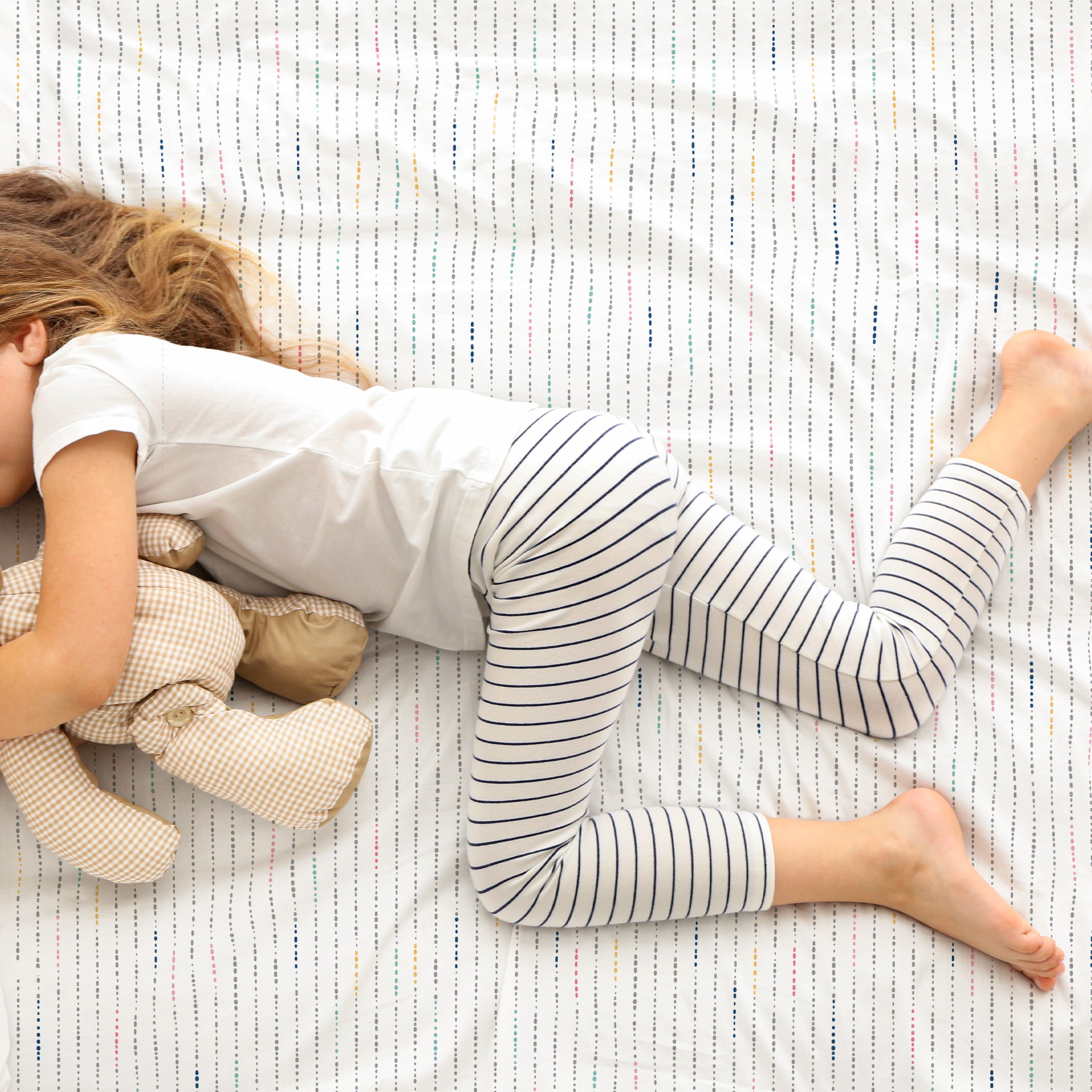 A young child in striped pajamas lying face down on a bed with white and red striped Organic Cotton Sheet Set - Pebble Pop sheets, hugging a teddy bear.