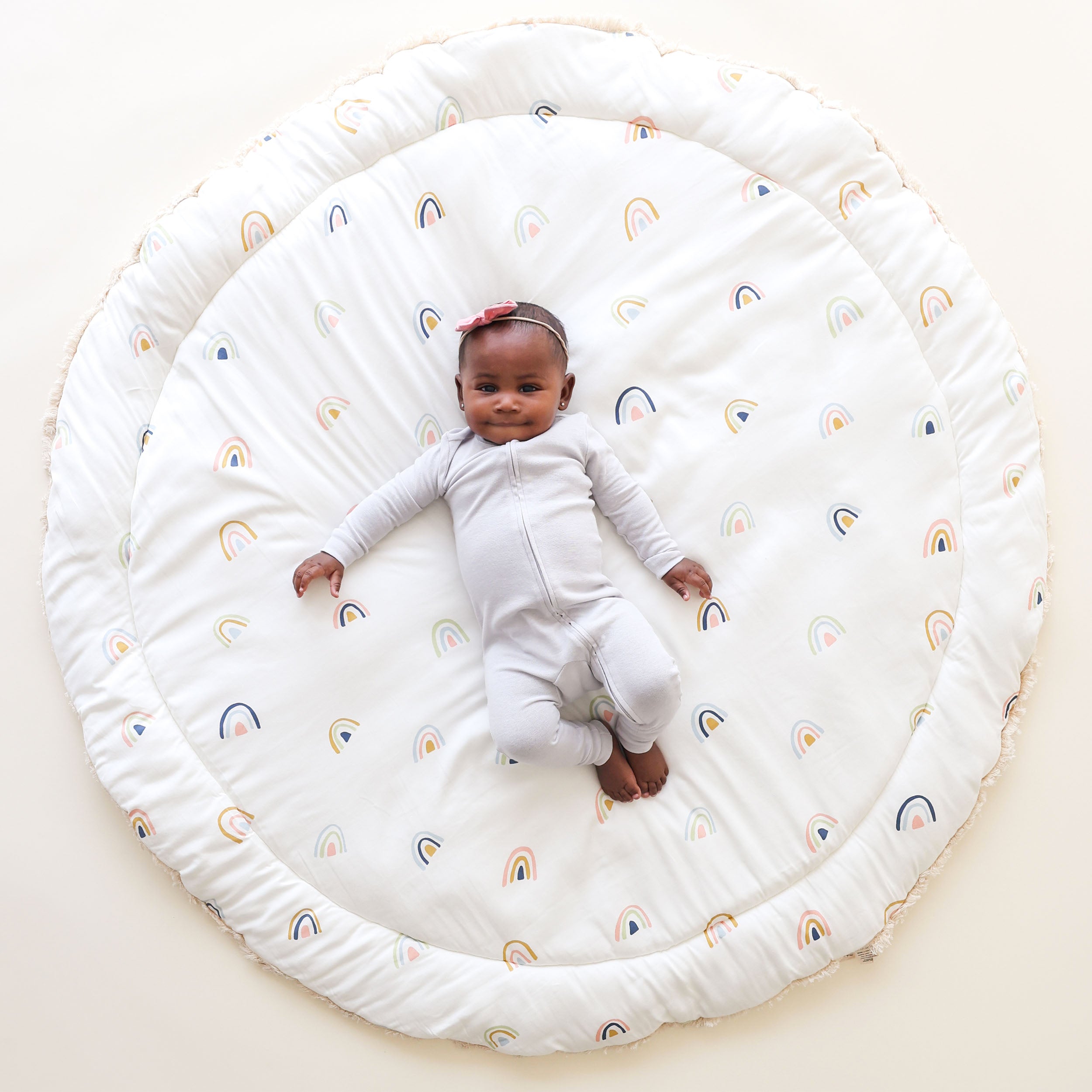 A cheerful baby with a red headband lies on a Makemake Organics Organic Cotton Quilted Reversible Play Mat adorned with colorful rainbows. The baby wears a white bodysuit and has a joyful expression.