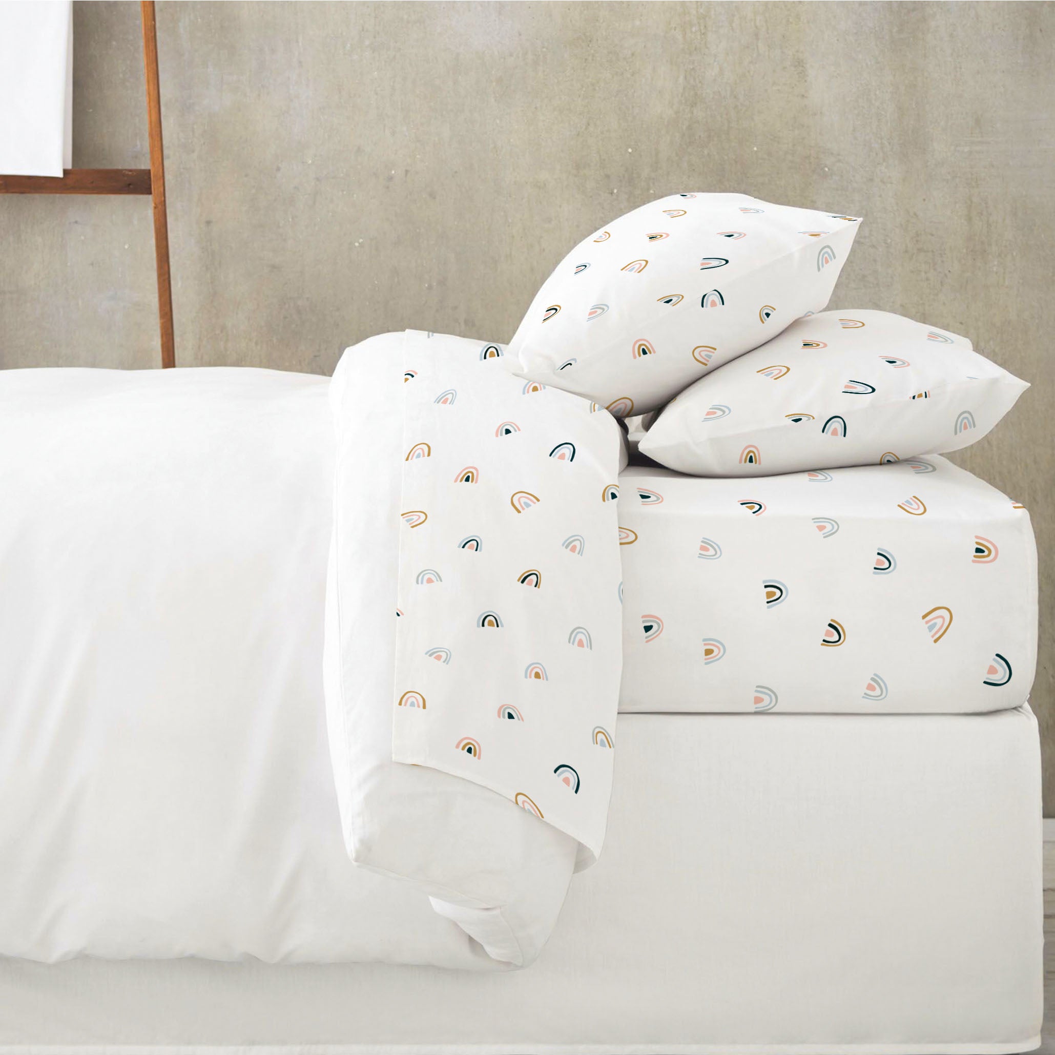 A neatly made bed featuring a white duvet and pillows with a playful Over The Rainbow organic cotton sheet set cover in a room with a concrete wall by Makemake Organics.