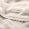 Close-up of a cream-colored Organic Cotton Scalloped Baby Blanket showing detailed texture and a brand label that reads "Makemake Organics".