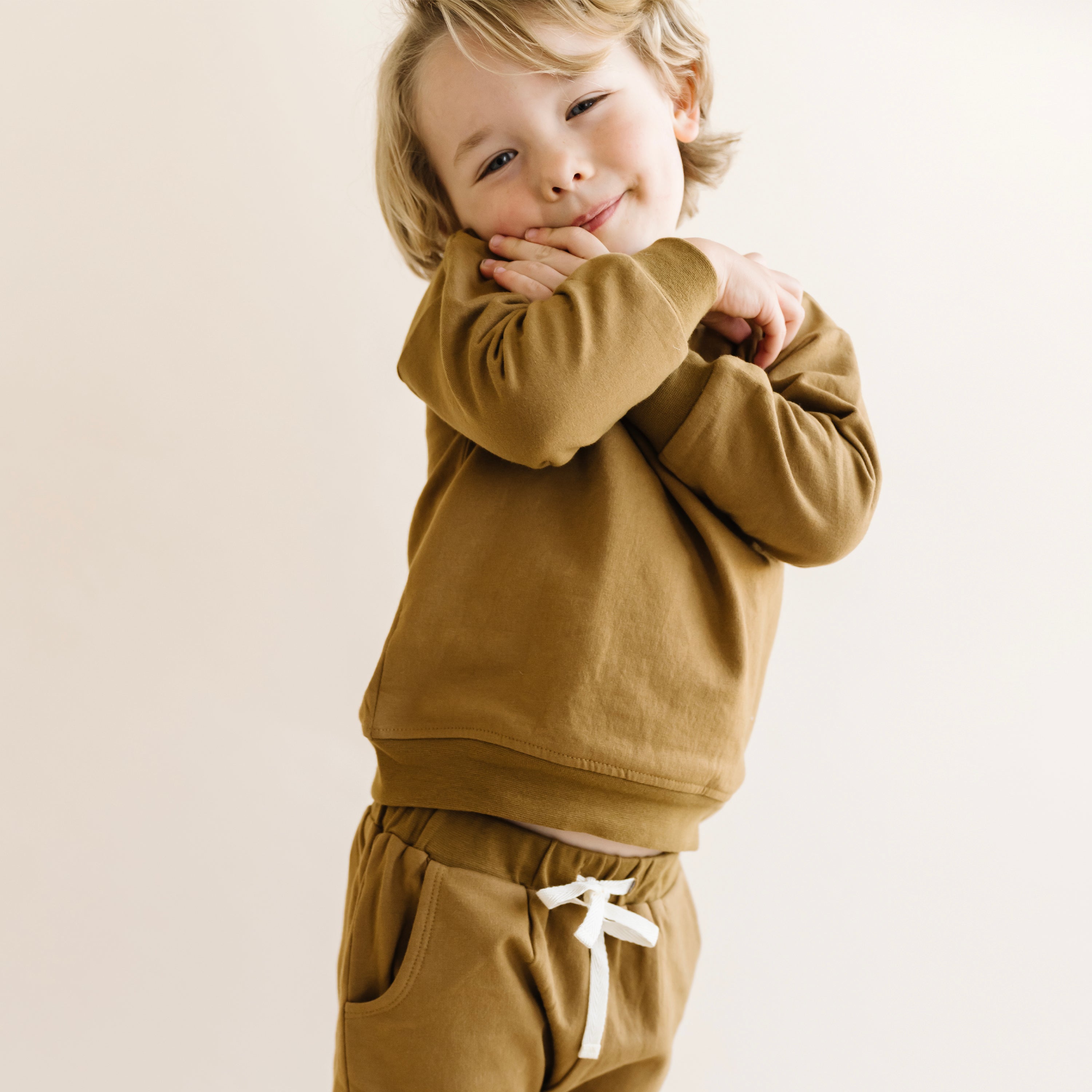 A young child wearing a cozy brown Organic Sweatshirt - Walnut Solid and matching pants from Organic Kids smiles charmingly with hands clasped under their chin, standing against a light beige background.