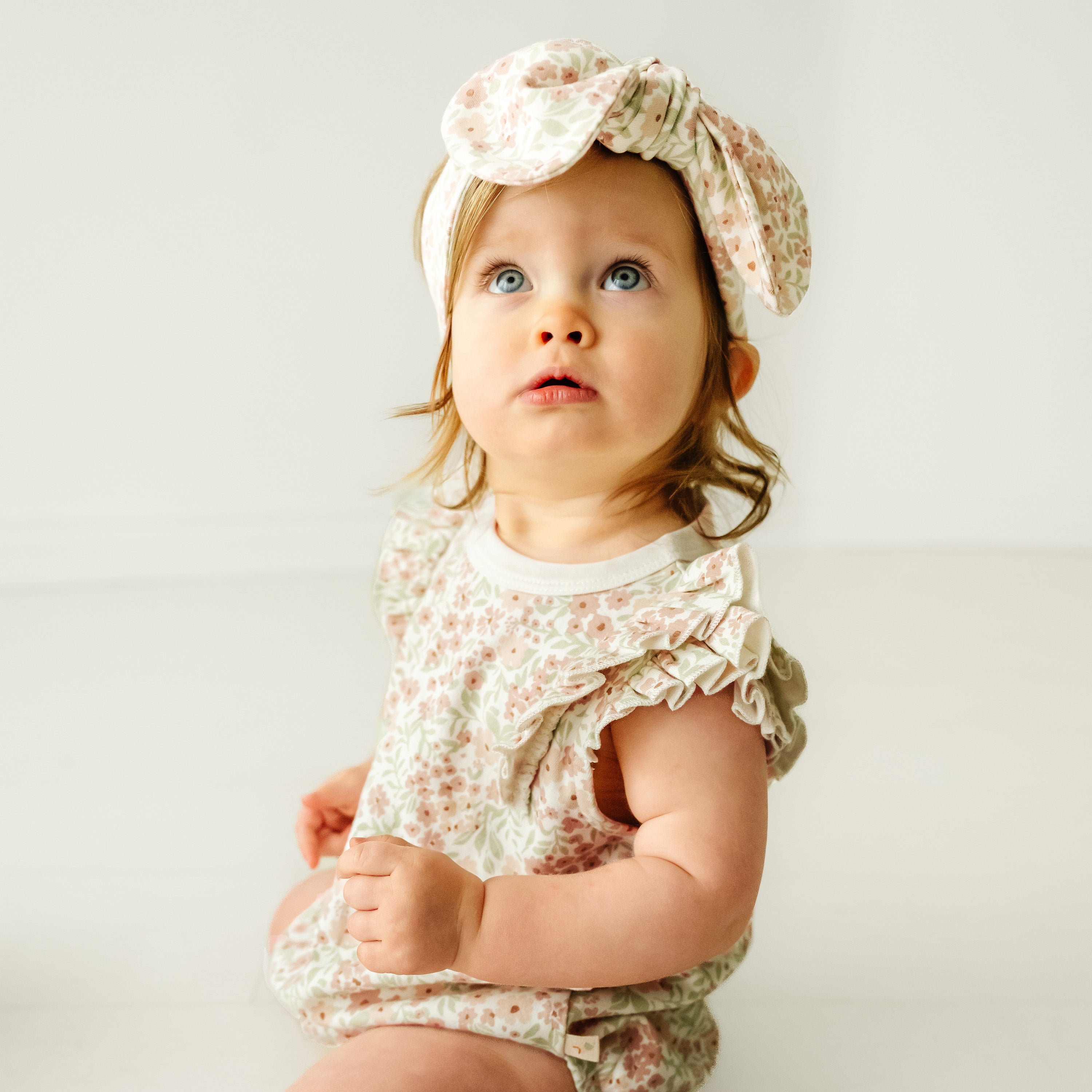 A baby with wide blue eyes and light hair, wearing a Makemake Organics Organic Flutter Bubble Onesie in Summer Floral and a matching bow headband, sits against a white background and looks upwards.