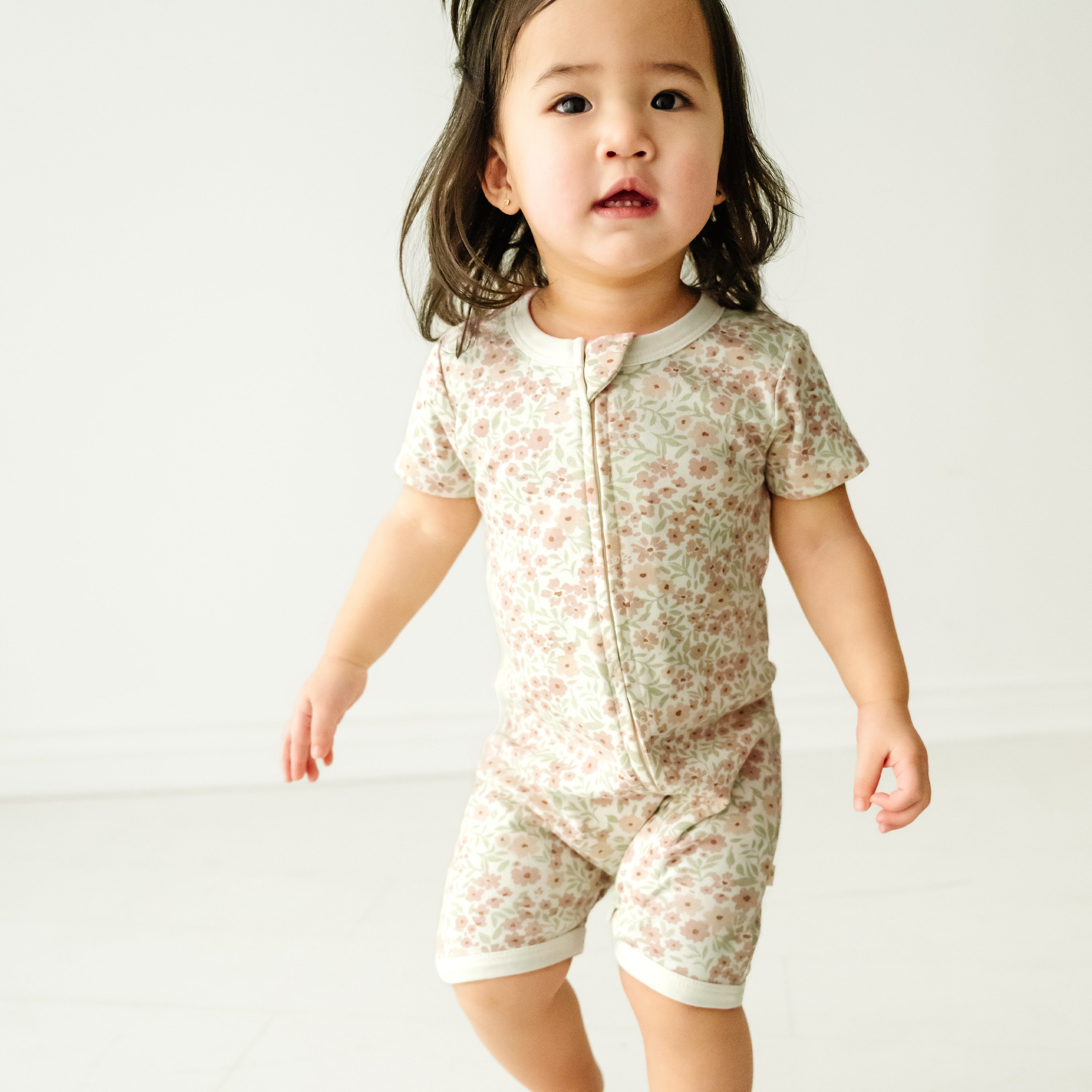 A toddler girl in a Makemake Organics Organic Short Zip Romper - Summer Floral standing in a bright room with a plain white background, looking slightly to the side with a thoughtful expression.