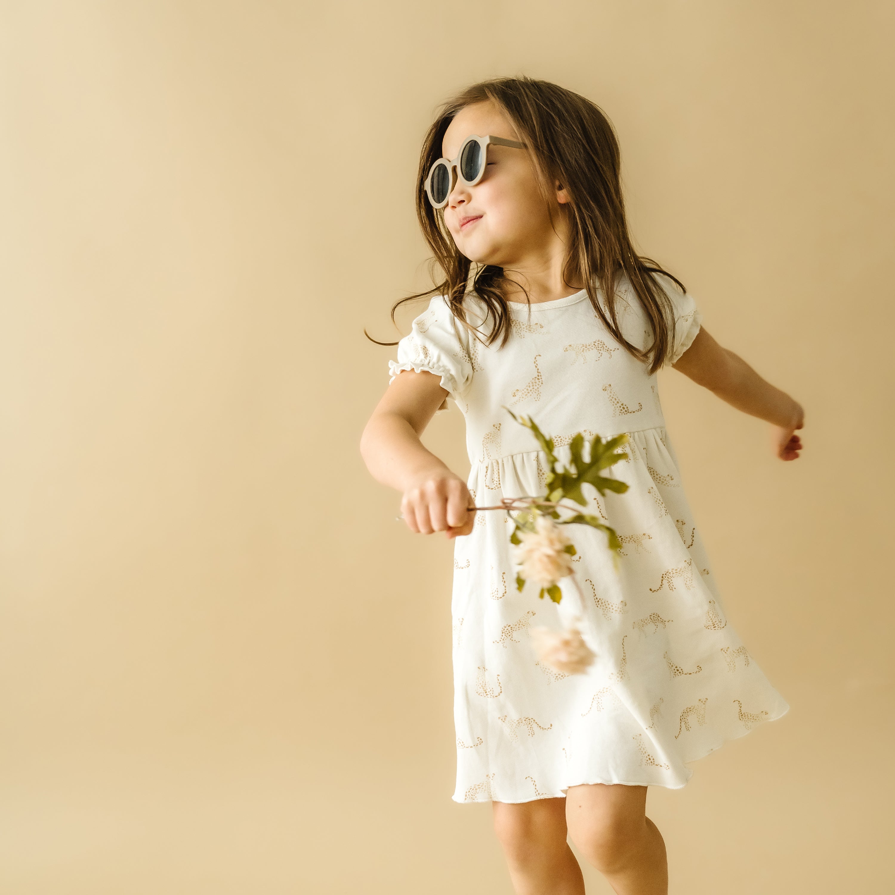 A young girl in a white printed Organic Kids Wildcat Puff Sleeve Dress and oversized sunglasses happily dances, holding a twig with leaves. She's posing against a light beige background.