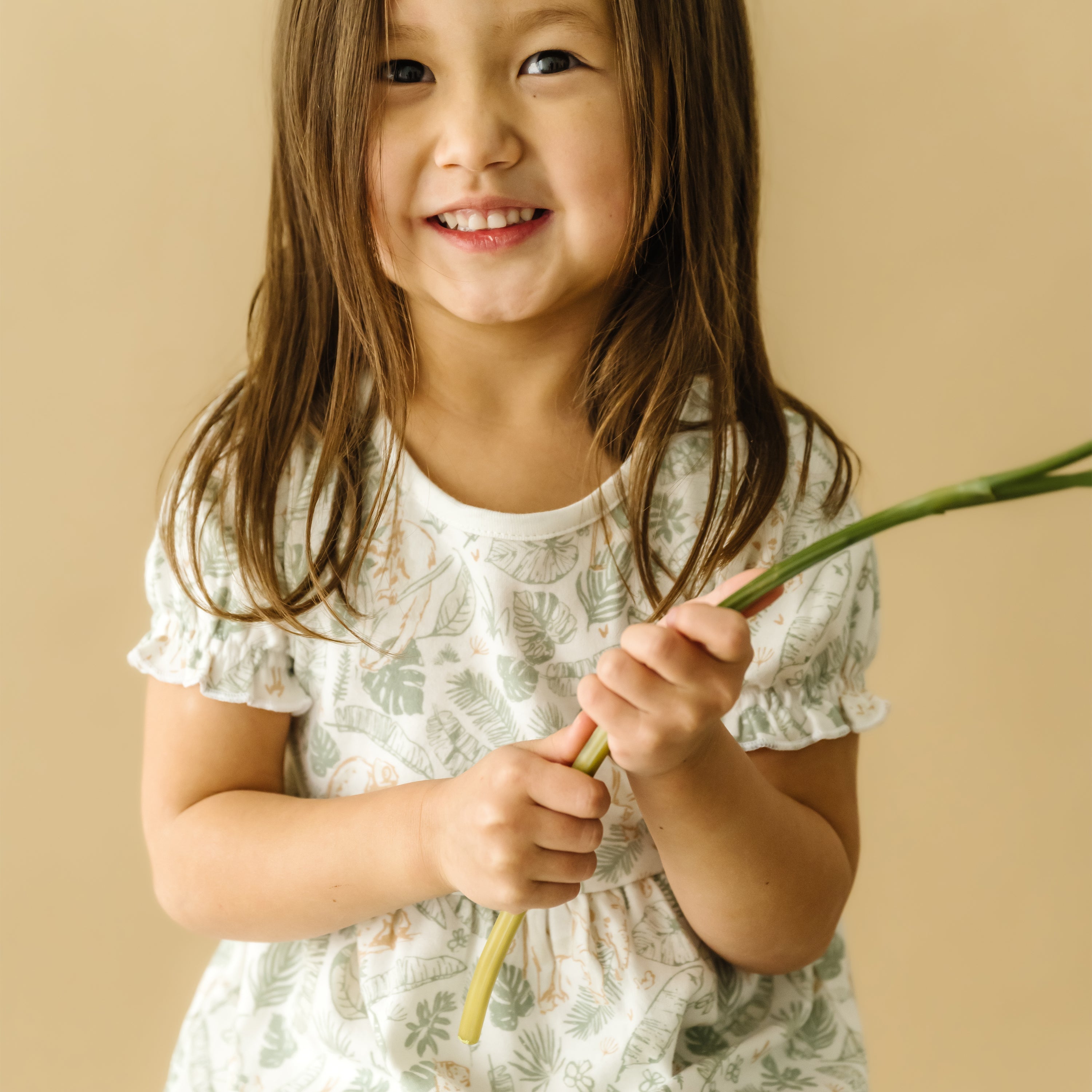 A young girl with long hair, smiling, holding a green flower stem from Makemake Organics, wearing a Organic Puff Sleeve Dress - Wild Safari by Organic Kids, against a light beige background.