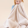 A young child with red hair peeking over a large, soft Organic Cotton Scalloped Baby Blanket - Nora Shell draped around them, set against a warm beige background by Makemake Organics.