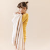 A young girl stands against a soft beige background, holding an Organic Cotton Scalloped Baby Blanket in Nora Shell from Makemake Organics. she wears a yellow long-sleeve shirt and looks thoughtfully to the side.