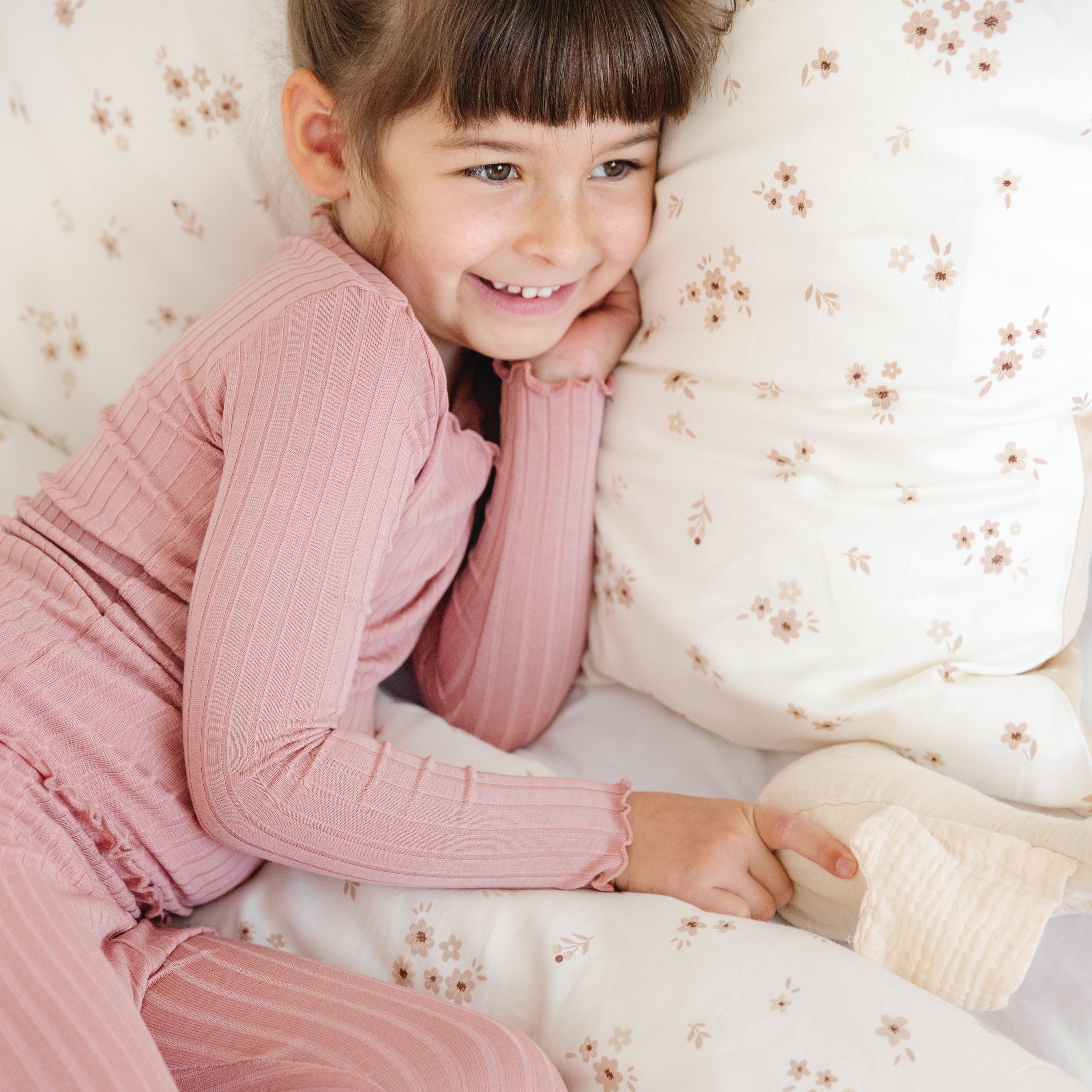 A young girl in pink pajamas smiles while lying on a Makemake Organics Organic Cotton Sheet Set - Bloom, holding onto a small, soft toy.
