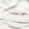 Close-up of a white knitted fabric with a "Makemake Organics" label, displaying textured knit patterns and folded layers of the Organic Cotton Scalloped Baby Blanket - Ella Ivory.