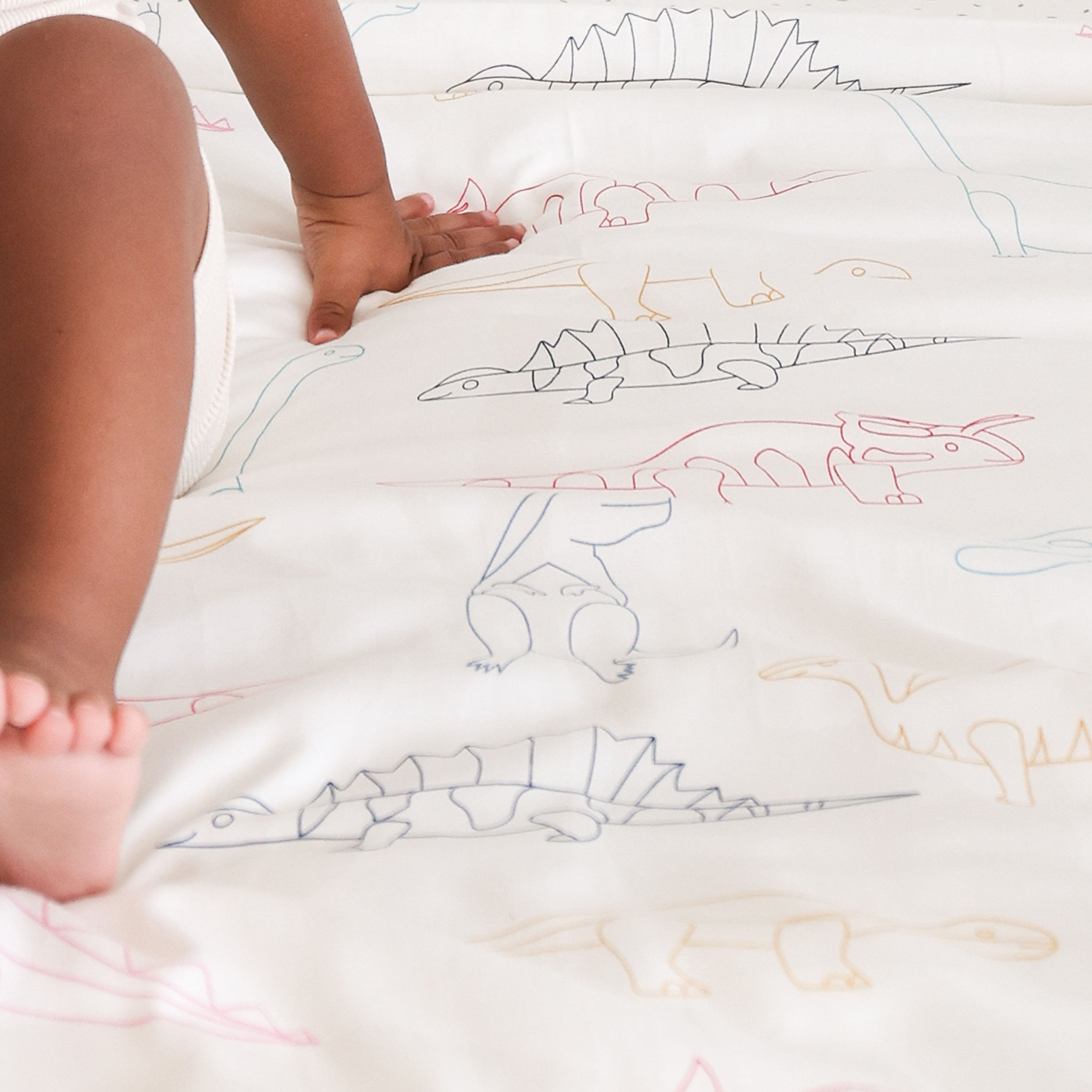 A toddler's bare feet on a bed with a Dino Park crib fitted sheet and pillowcase from Makemake Organics, featuring colorful outlines of various dinosaurs.