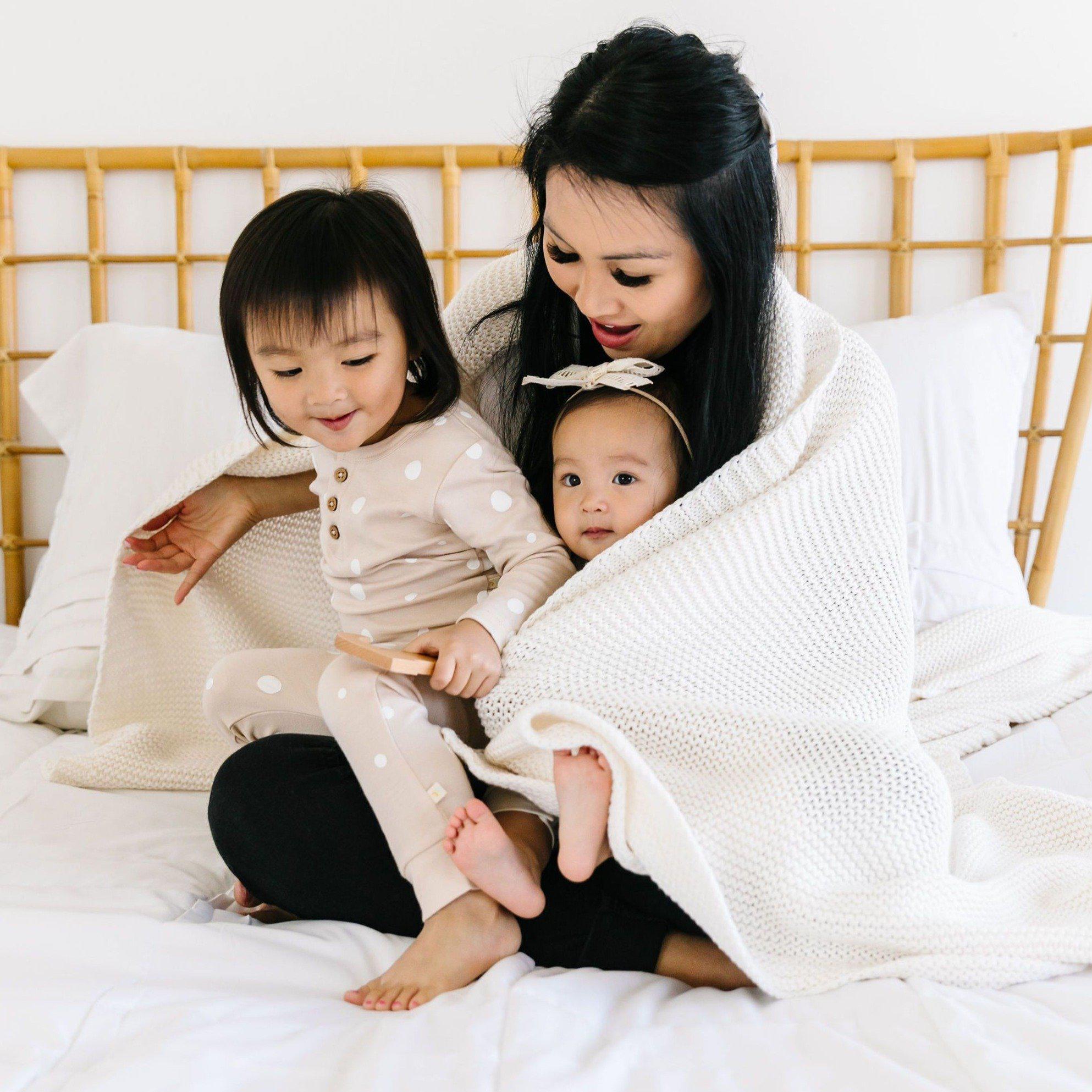 A mother and her two young children wearing pajamas are sitting on a bed, covered with a Chunky Knit Throw Blanket - Ella Ivory from Makemake Organics, smiling and enjoying playtime together.
