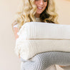 A smiling woman with blonde curly hair holding a stack of three cozy, Ella Ivory Chunky Knit Throw Blankets in neutral colors from Makemake Organics.