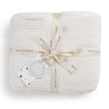 A neatly folded Ella Ivory Chunky Knit Throw Blanket made of organic cotton with subtle texture, tied with a ribbon and care label around it, featuring the brand name "Makemake Organics." Decorative leaf motifs are printed on the ribbon.