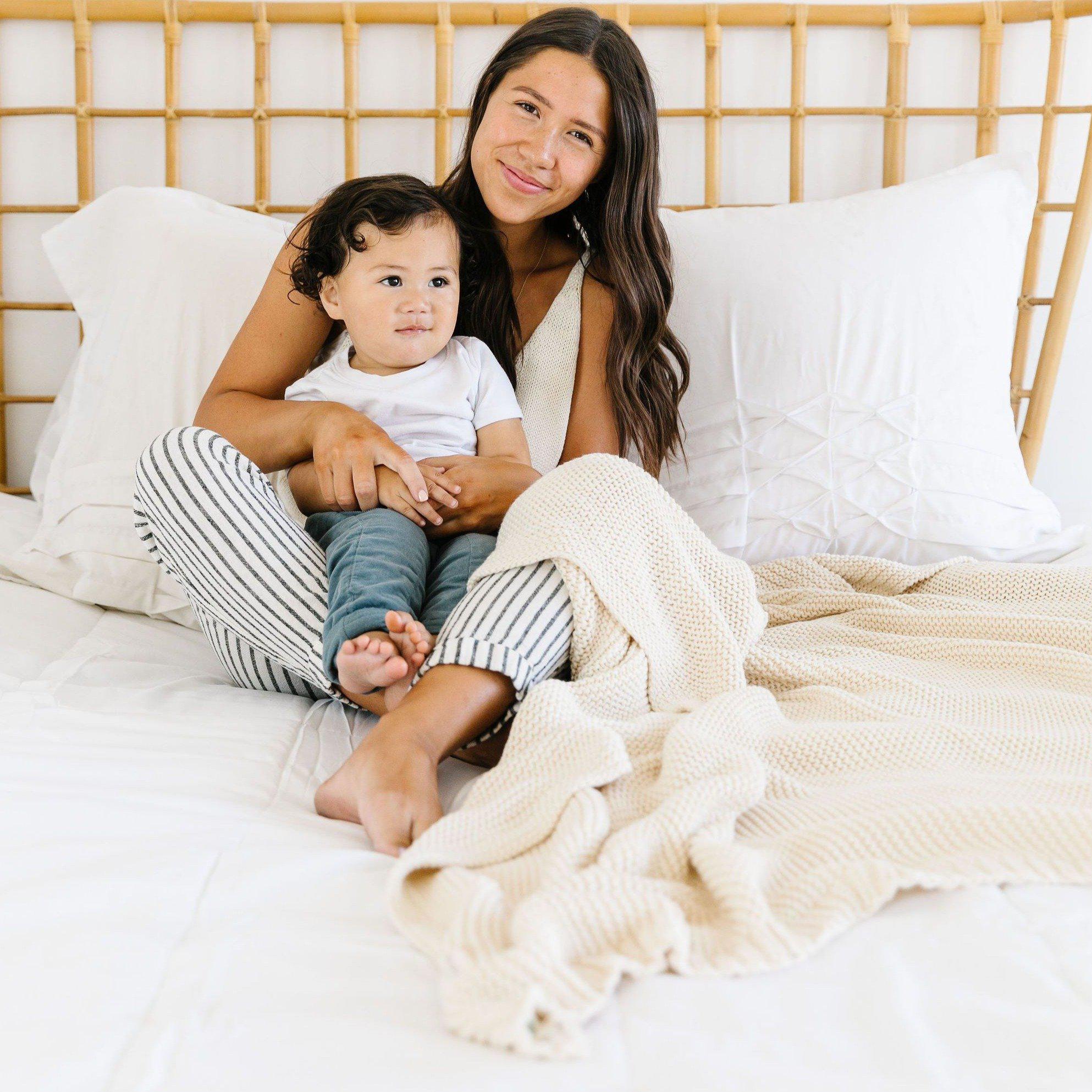 A mother and her baby sitting on a bed, surrounded by pillows, smiling at the camera. The mother is leaning against a rattan headboard, and a Makemake Organics Chunky Knit Throw Blanket - Vanilla Natural is nearby.