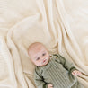 A baby in a green knit sweater lies on a Chunky Knit Throw Blanket - Vanilla Natural from Makemake Organics, looking upwards with a curious expression.