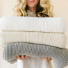 A woman holding a stack of three Makemake Organics Chunky Knit Throw Blankets in cream, beige, and gray colors, with only her lower face and hands visible.