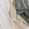 Close-up of three Chunky Knit Throw Blankets in neutral colors: white, beige, and gray, arranged in an overlapping design showcasing different patterns by Makemake Organics.