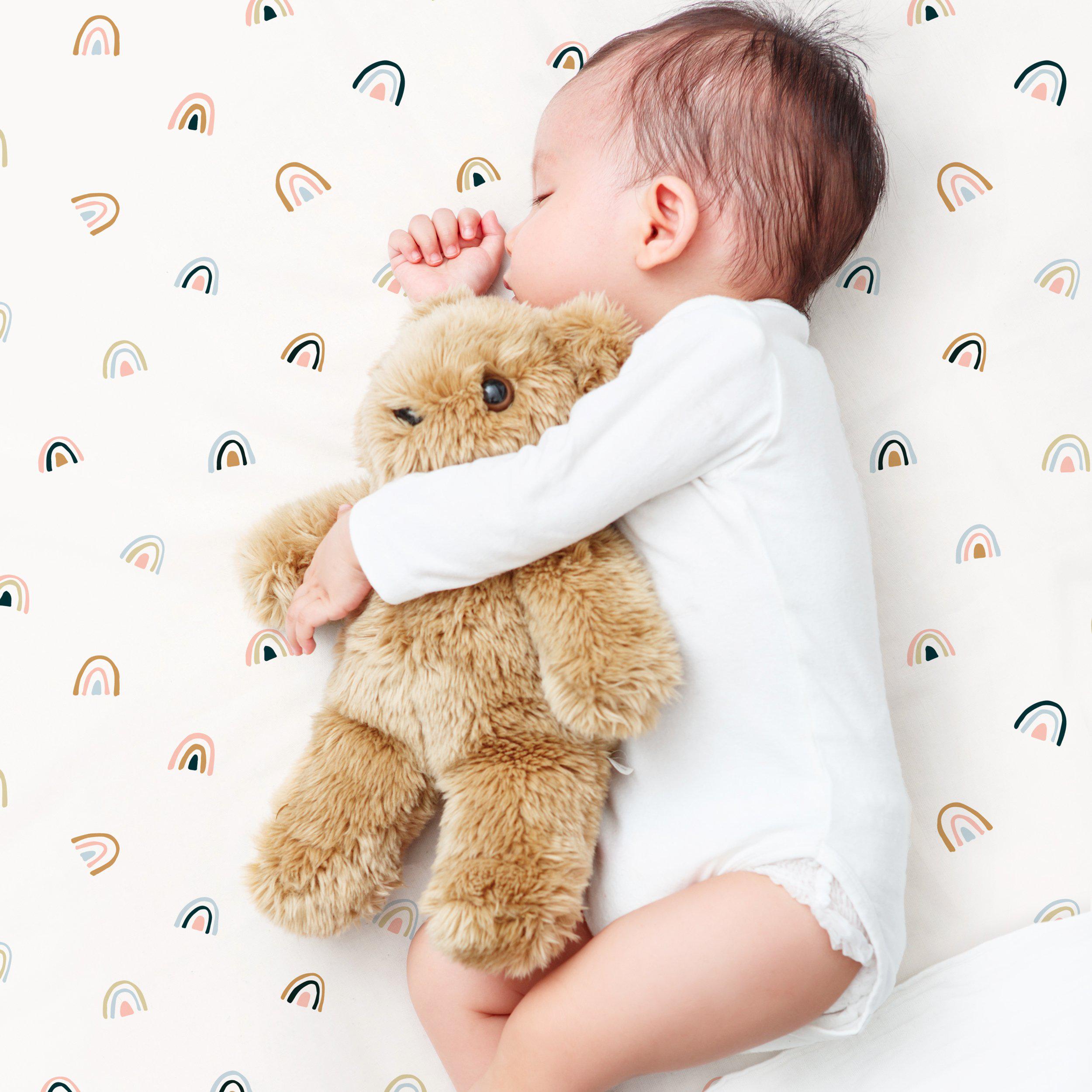 A baby peacefully sleeping while hugging a soft teddy bear, on a light background decorated with the Makemake Organics Crib Fitted Sheet with Pillowcase - Over The Rainbow.