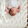 A newborn baby sleeps peacefully on a Celestial crib fitted sheet with pillowcase from Makemake Organics, featuring space-themed designs including rockets and planets. A white stuffed bunny toy is partially draped over the baby.