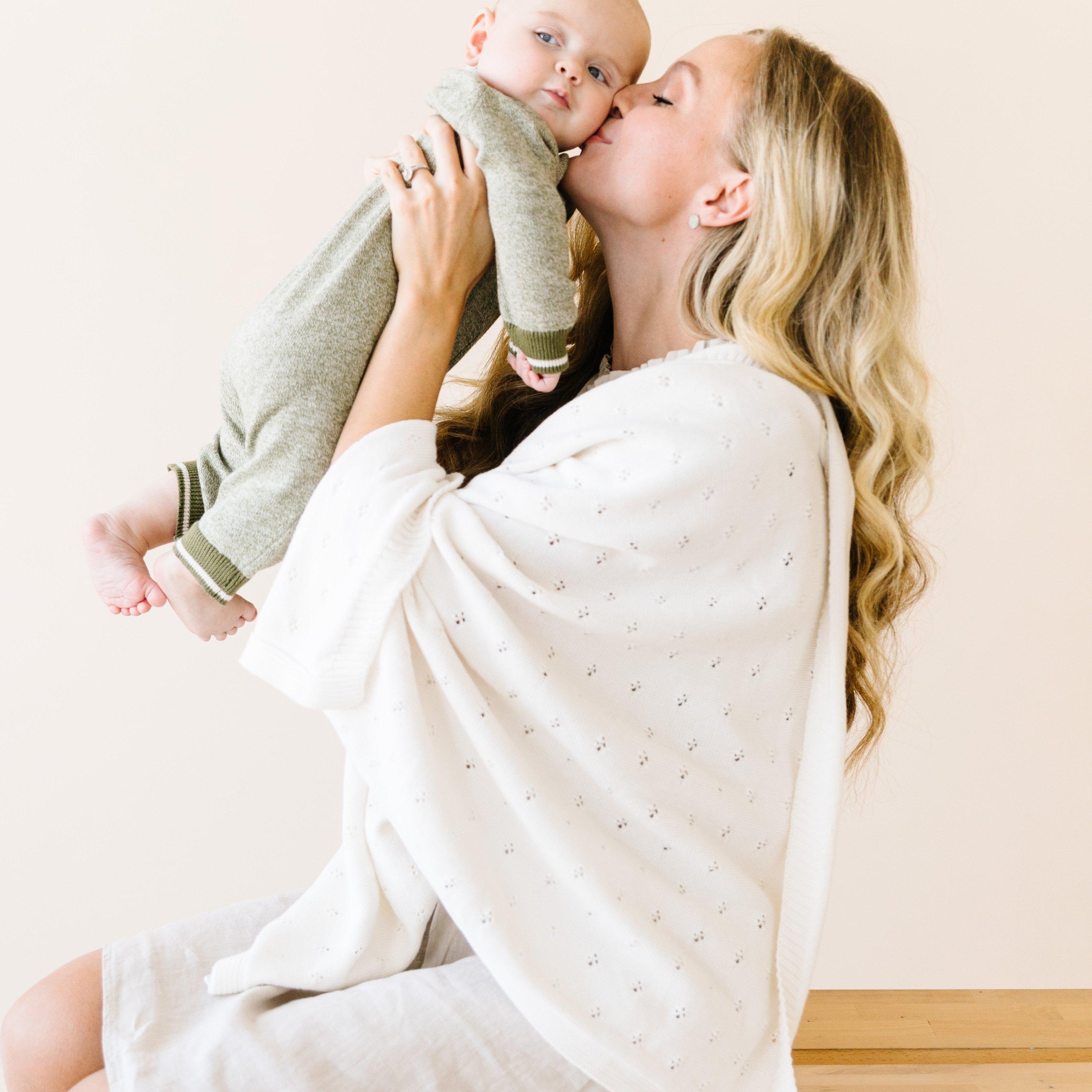 A mother lovingly kisses her baby, holding them in her arms against a soft pink background, both dressed in light, soft colors with the Makemake Organics Organic Cotton Pointelle Baby Blanket - Ella Ivory.