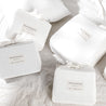 Four white, square cosmetic packages with "Makemake Organics skincare" labels, beautifully tied with ribbons, arranged neatly on a soft, crumpled linen background.