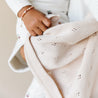 A close-up image of a baby's small hand holding a Makemake Organics Organic Cotton Pointelle Baby Blanket in Nora Shell with delicate floral patterns. The cozy fabric suggests warmth and comfort.