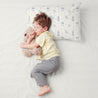 A young boy sleeps peacefully on a white bed, hugging a teddy bear, with a Makemake Organics Organic Cotton Toddler Pillowcase - Celestial adorned with rocket ship patterns next to him. he is wearing a yellow t-shirt and gray pants.