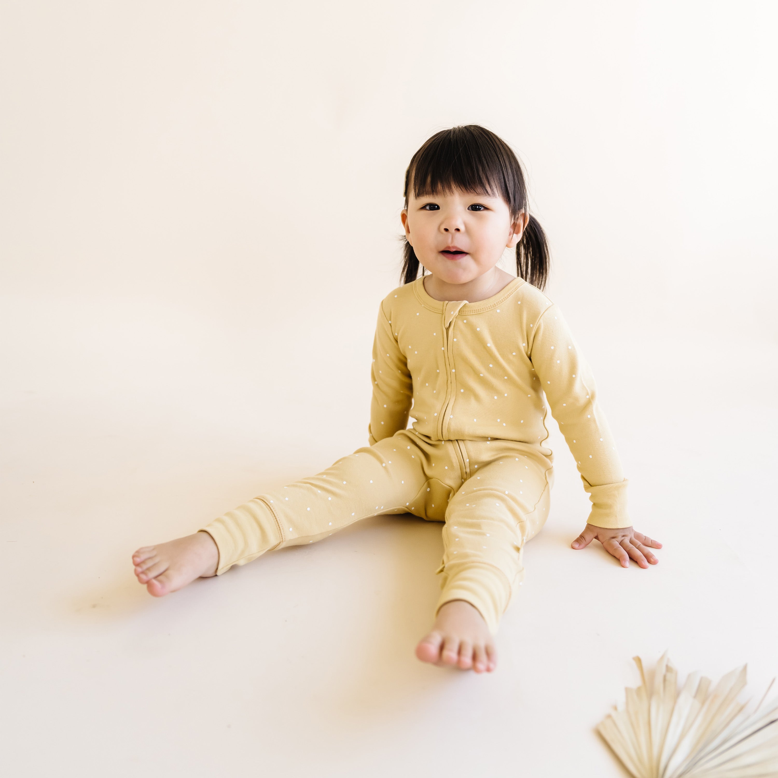 A toddler with short black hair, wearing an Organic Baby Organic 2-Way Zip Romper - Yellowstone with a polka dot pattern, sits on a pale background, looking at the camera with a neutral expression.