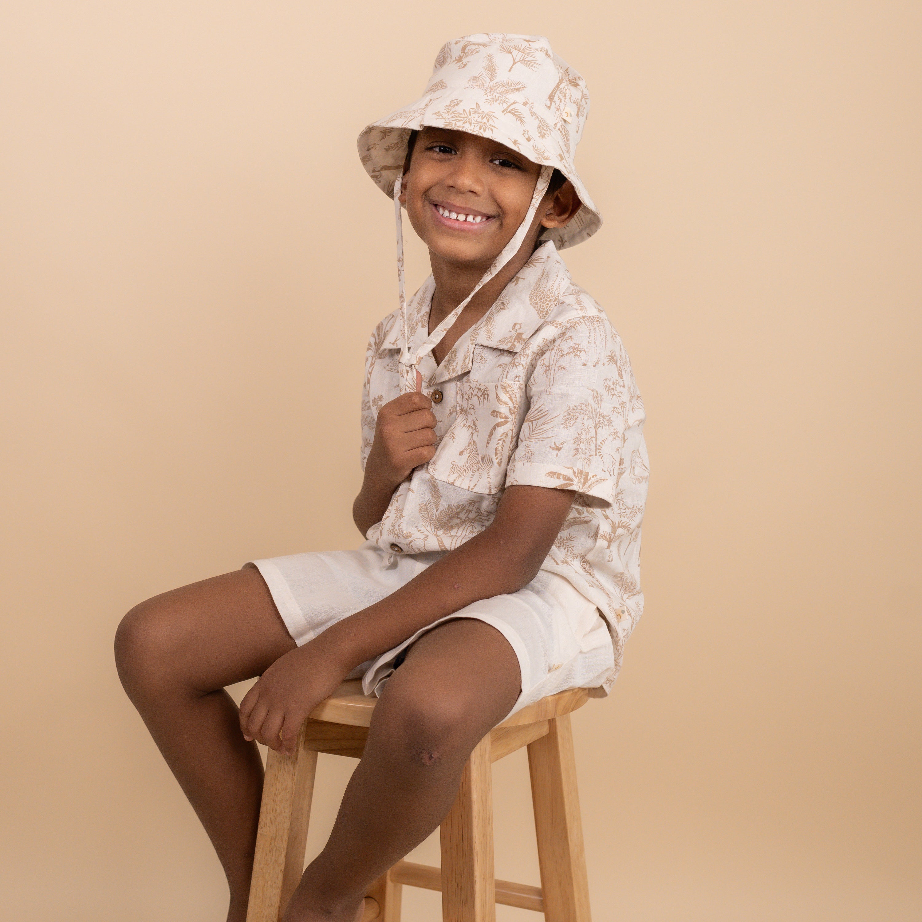 A smiling child wearing a floral bucket hat and matching shirt sits on a wooden stool against a beige background. The outfit features light colors with a subtle pattern.
