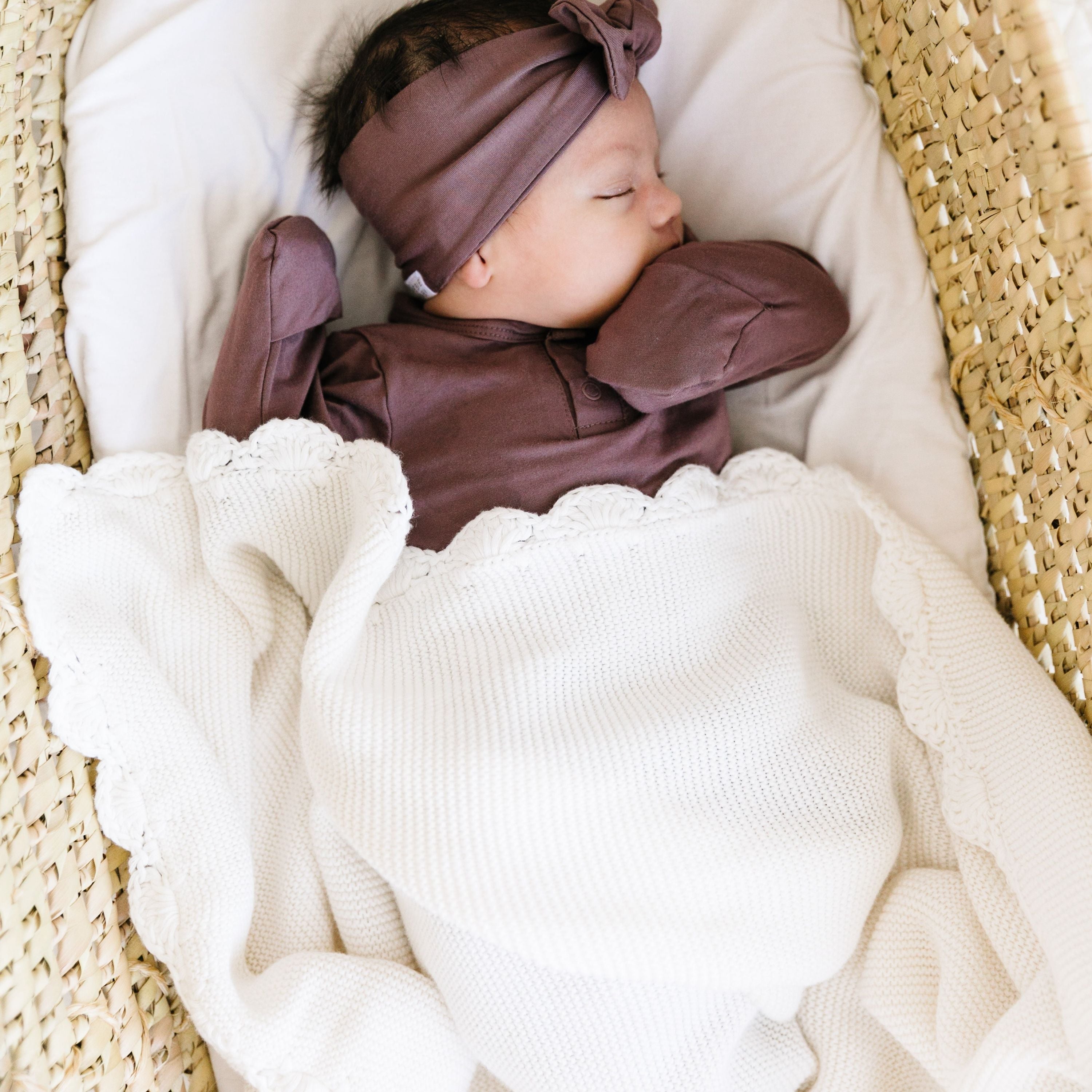 Baby sleeping in a woven basket, wearing a purple outfit and headband, with a white lace-trimmed blanket.