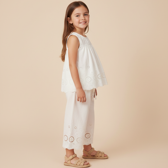 A young girl with long brown hair stands sideways, smiling, wearing a sleeveless white top and matching white pants with eyelet details, and beige sandals, against a beige background.